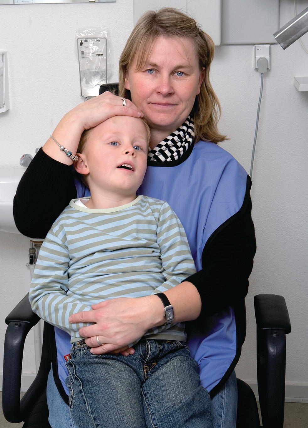 Photo of a woman (parent) with a child placed on her lap, holding the child’s hand with one hand and support the child’s head with the other hand.